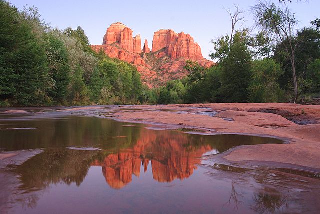 Photograph of red rocks in Arizona overlooking a pool of water ringed with evergreen trees. It is evening.