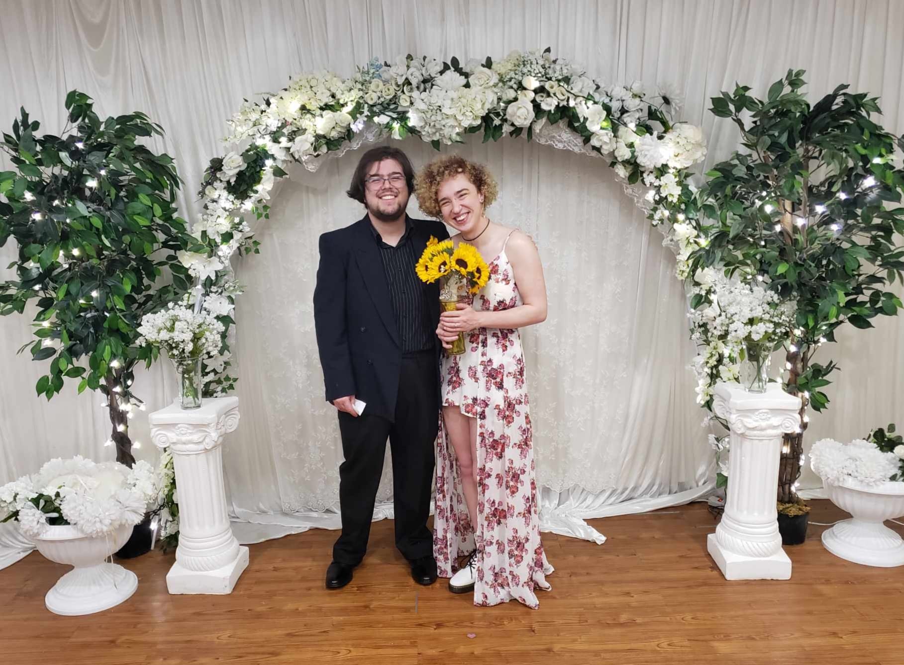 Demitri and I in front of a wedding arch. I am holding a bouquet of sunflowers