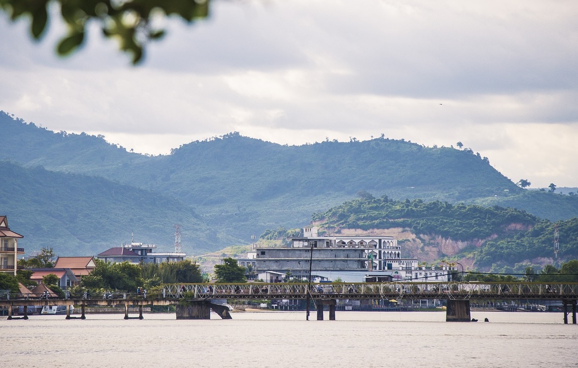 A photo of a scenic building, taken in Cambodia. There is ocean in the foreground, and rolling hills behind. There are some other smaller buildings nearby.