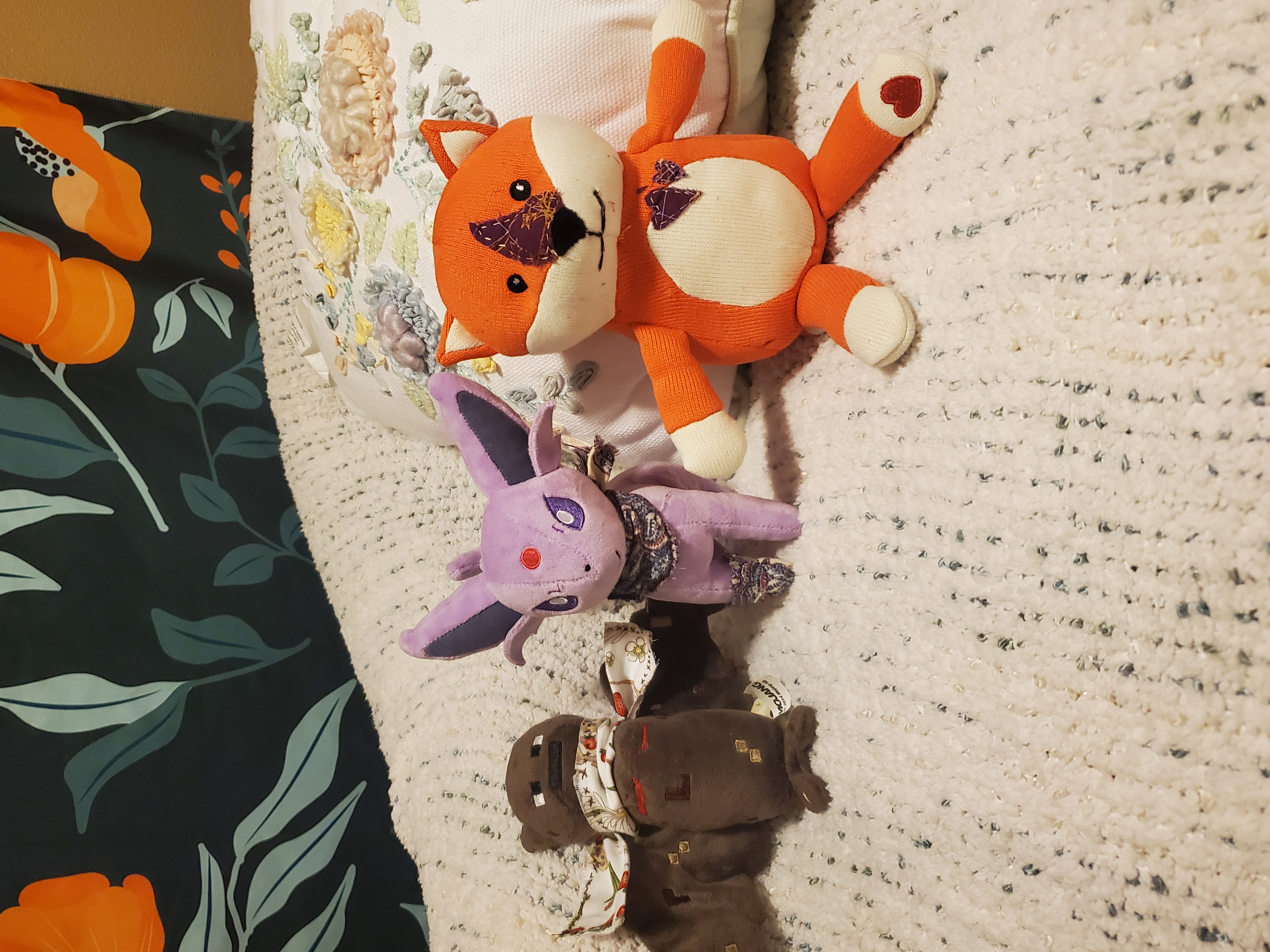 photo of 3 plushies. from left to right, first is a minecraft bat plushie from 2014 with a cross-sitched scarf, patterned with strawberries and mice. it also has embroidered top surgey scars sown on. second is an espeon plushie with a purple, white and magenta scarf and sock. third plushie is a bright orange, lopsided fox plushie with embroidered black eyes and nose. they also white paws, belly, cheeks, and ears with a red heart on their right foot. their snout has purple fabric stitched on, along with purple hearts on their belly. they have black thread stitched on their face in a smile.