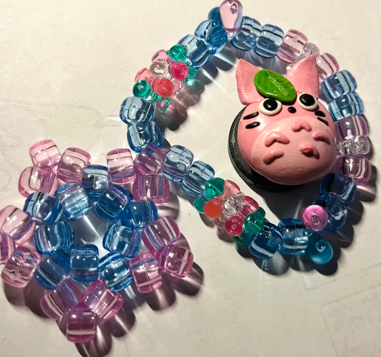 Two handcrafted items by Swamp God; a Kandi bracelet with trans pride colors and a beaded star, and a tiny magnet of a sakura-themed Totoro.