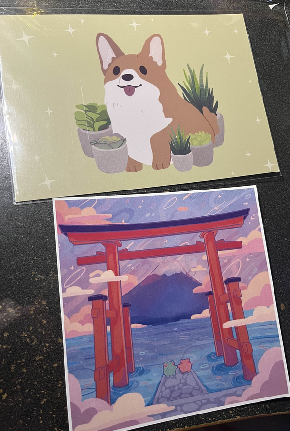 Small art prints illustrated by mussyhead and KimchiKrafts. Mussyhead's piece is of a cartoon corgi surrounded by succulents, and KimchiKrafts' piece is of two little froggies chilling under a big torii, with a majestic mountain as a backdrop.