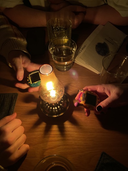 hand holding a tamagotchi v3 next to a small gas light on a table at a dark bar