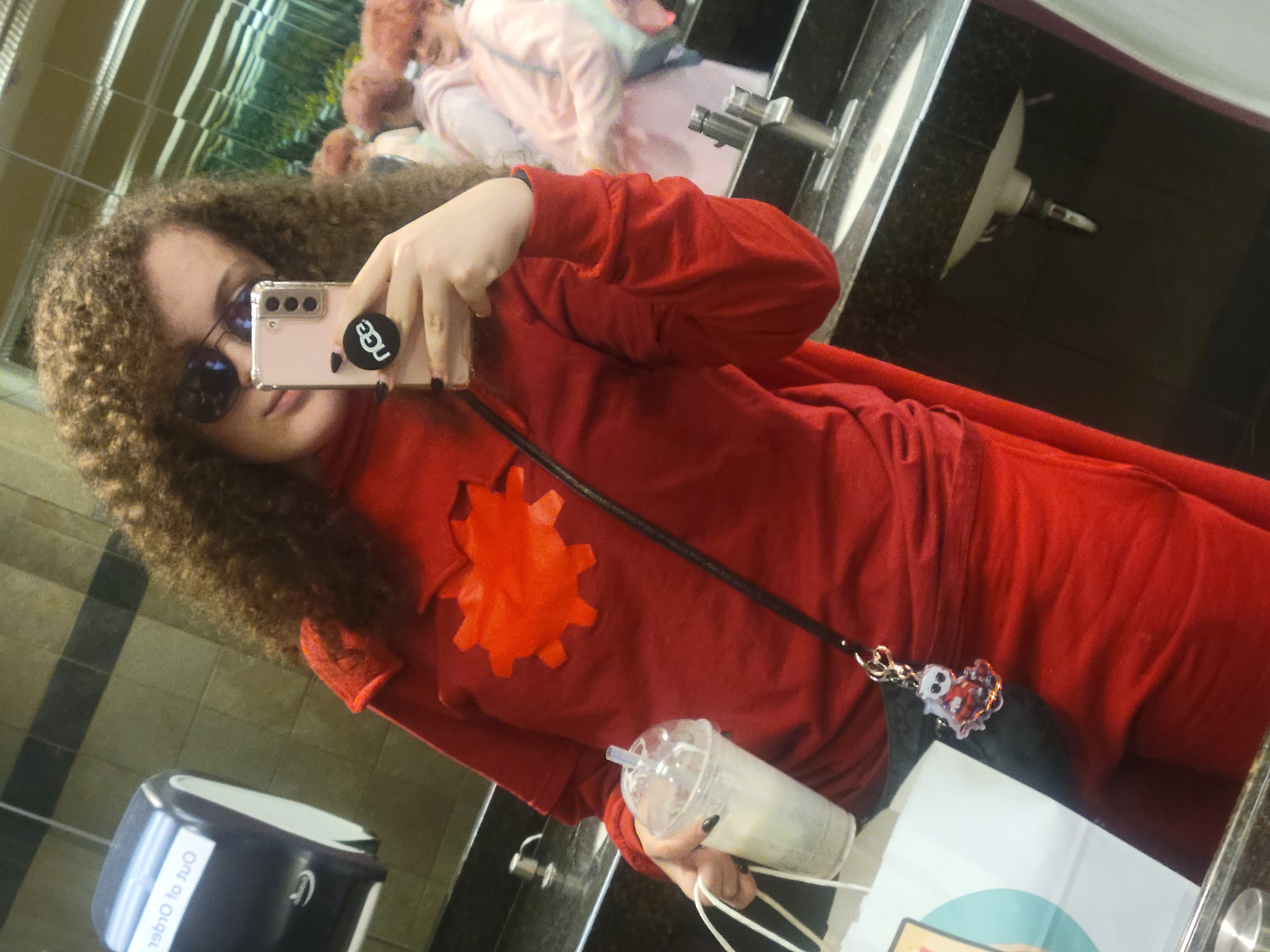 a woman with pale skin takes a mirror selfie in a public bathroom. he's dressed up in dave strider's knight of time uniform. the hood is down, revealing long brunette curly hair. he wears sunglasses on his face. theres a purse with davekat charms attached and in his other hand he holds a bag and a drink.