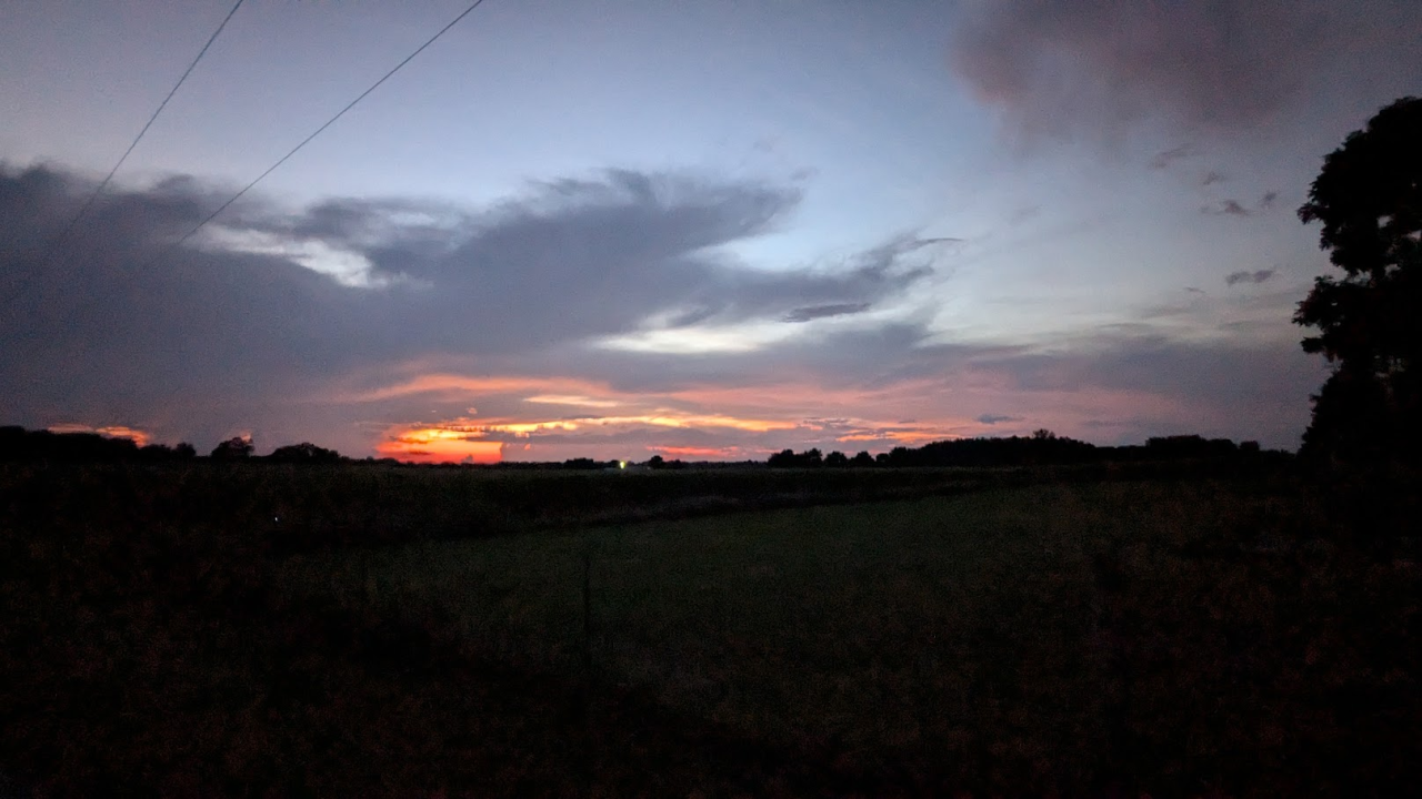 A slightly fuzzy picture of dusk. It's so dark that the treeline is completely black. Violent red-to-yellow reflects off of grey clouds. Above the clouds, the sky is a very pale and slightly muted blue.