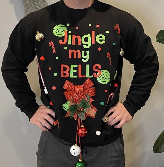 A person is standing square on to the camera with their hands on their hips wearing black jeans and black jumper. The jumper has red and green writing on it and candy cane and Christmas ornament motifs. Large printed letters read 'Jingle My Bells' across the front. Underneath the writing is a single red bow, with three sleigh bells hanging from ribbons. The lowest bell is level with the person's crotch.
