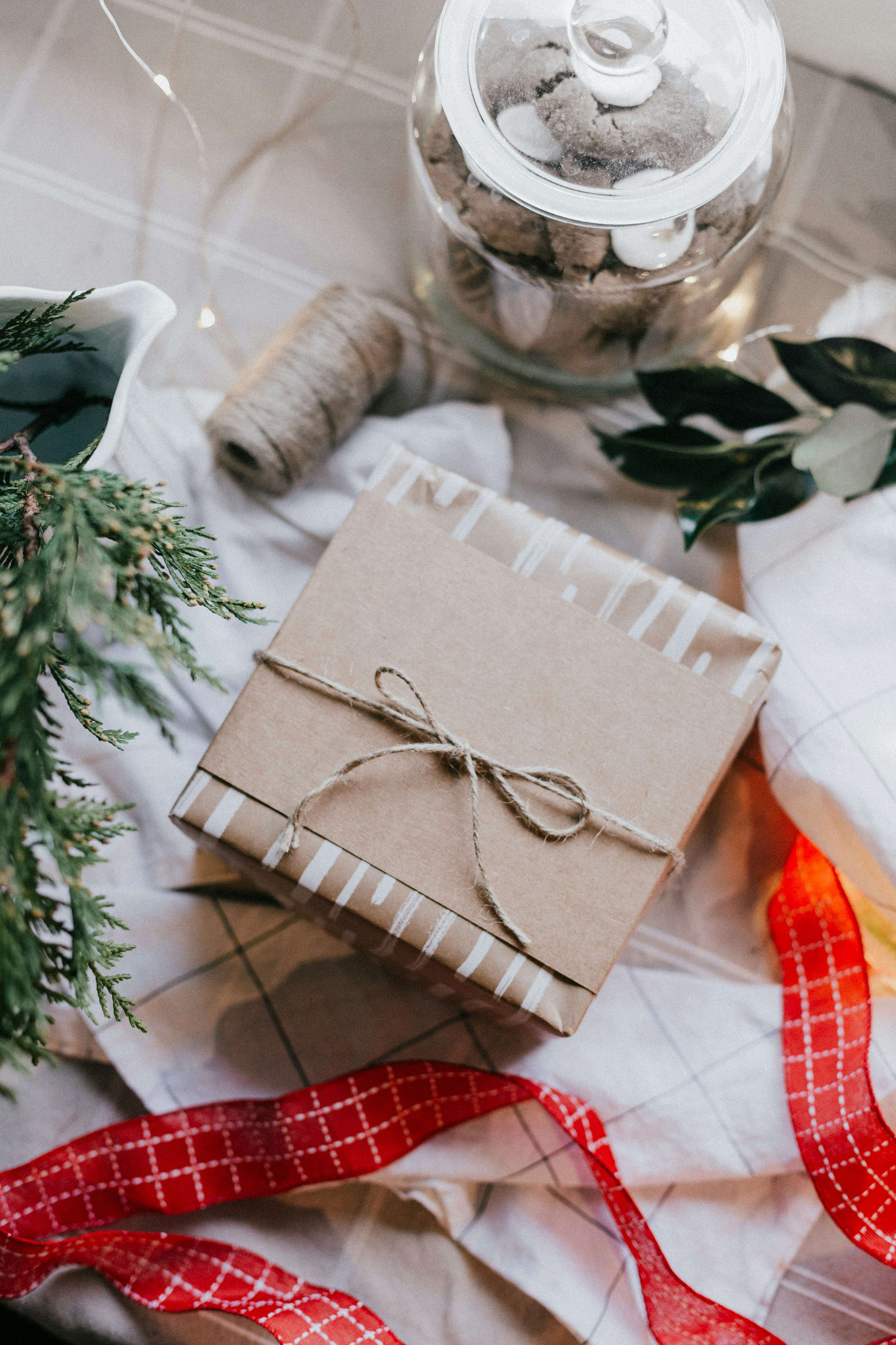 A single wrapped box is positioned on a table top and surrounded by gift-wrapping materials. The box is wrapped with irregularly striped gold and white paper, fastened with a band of brown paper and a piece of jute twine fashioned into a bow. The items surrounding the box include a small string of warm white fairy lights, a spool of twine, some red ribbon with gold checks, an evergreen frond, some leaves, and a clear glass jar of cookies.