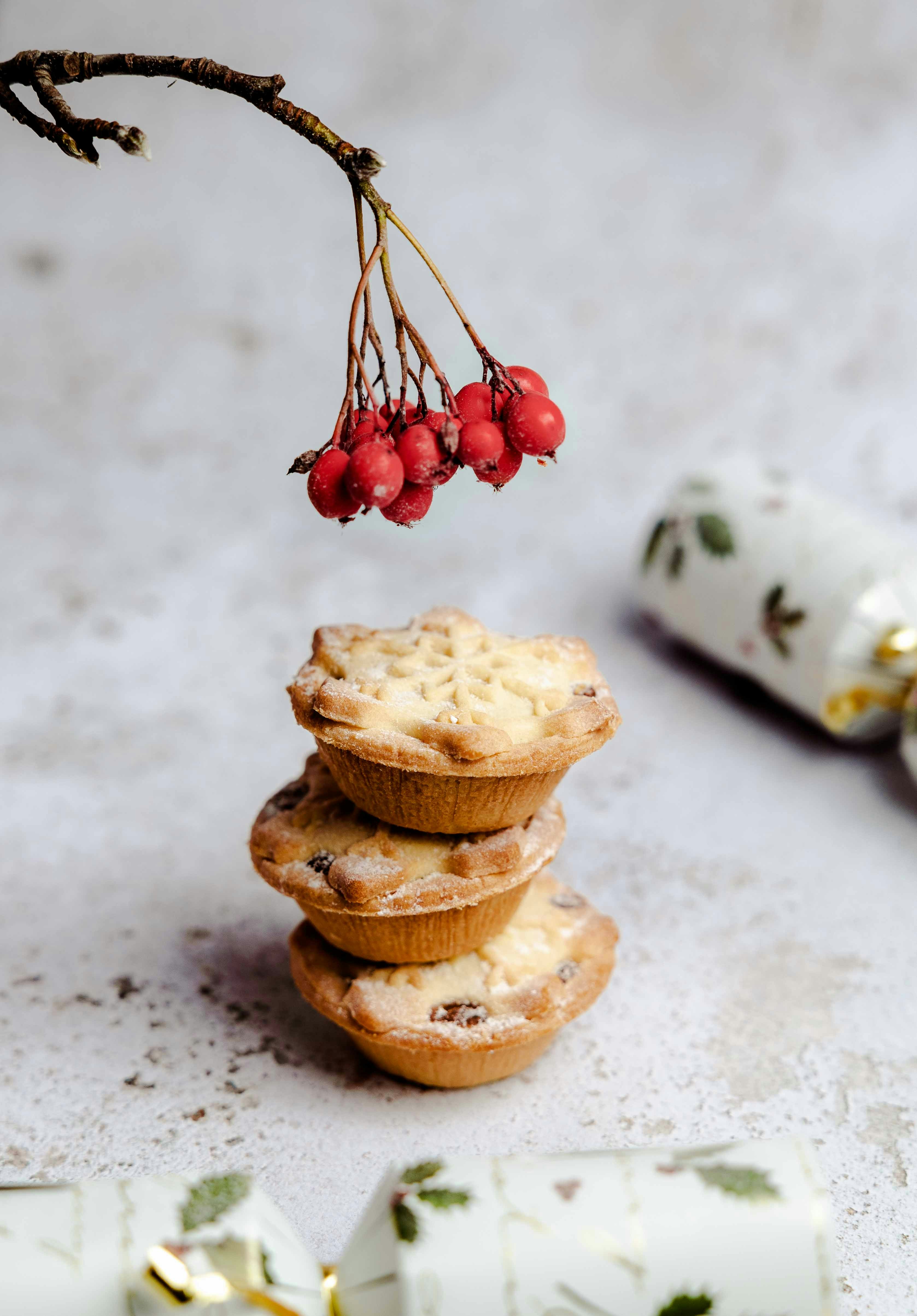A stack of three christmas mince pies sit on a table under a branch with a cluster of red berries at its tip.  The mince pies are decorated with snowflake impressions on the tops cut out of pastry. There are two white and gold Christmas crackers on the table. The table's surface is rough textured and pale, similar to formed concrete.
