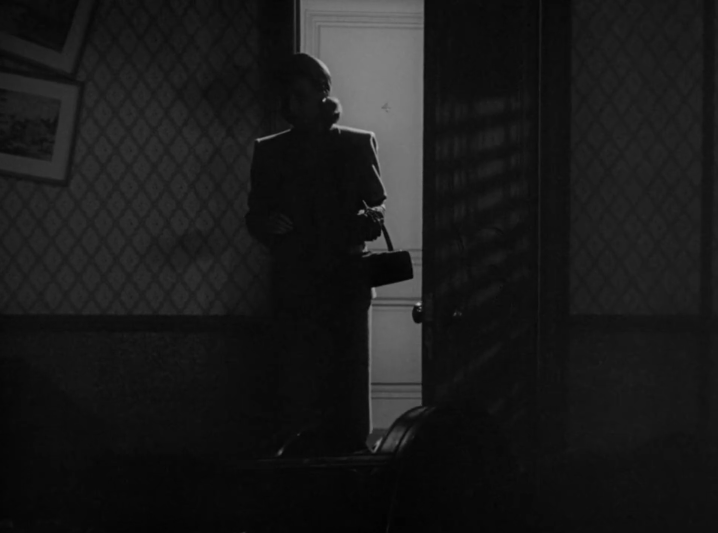 A screencap from Chicago Deadline. A feminine figure, her face cloaked in shadow, stands in a dark room only illuminated by the open door.