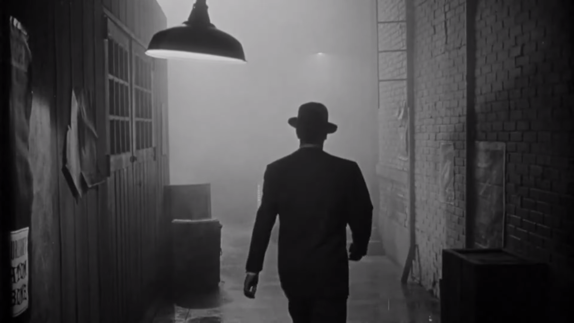 A screencap from The Big Combo. A man in a suit and hat, covered in shadow, walks through a foggy alley.