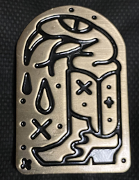 A metal pin with a black lined carved out design of a fanged snake sitting in a cowboy boot as venom drips from its fangs