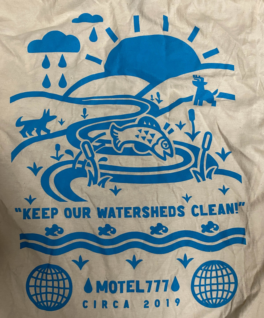 Close up of the design of a cream-colored t-shirt with a blue design showing a solid blocky happy nature scene with text that says keep our watersheds clean motel777 circa 2019