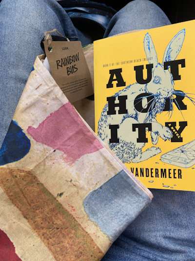 Photograph of the muted yet colorful knot wrap folded up in my lap with the book Authority sitting on it. The book is solid yellow with black text and the cover art is of a rabbit sitting next to a smart phone, the artwork's thin lines being a shining holographic sapphire blue