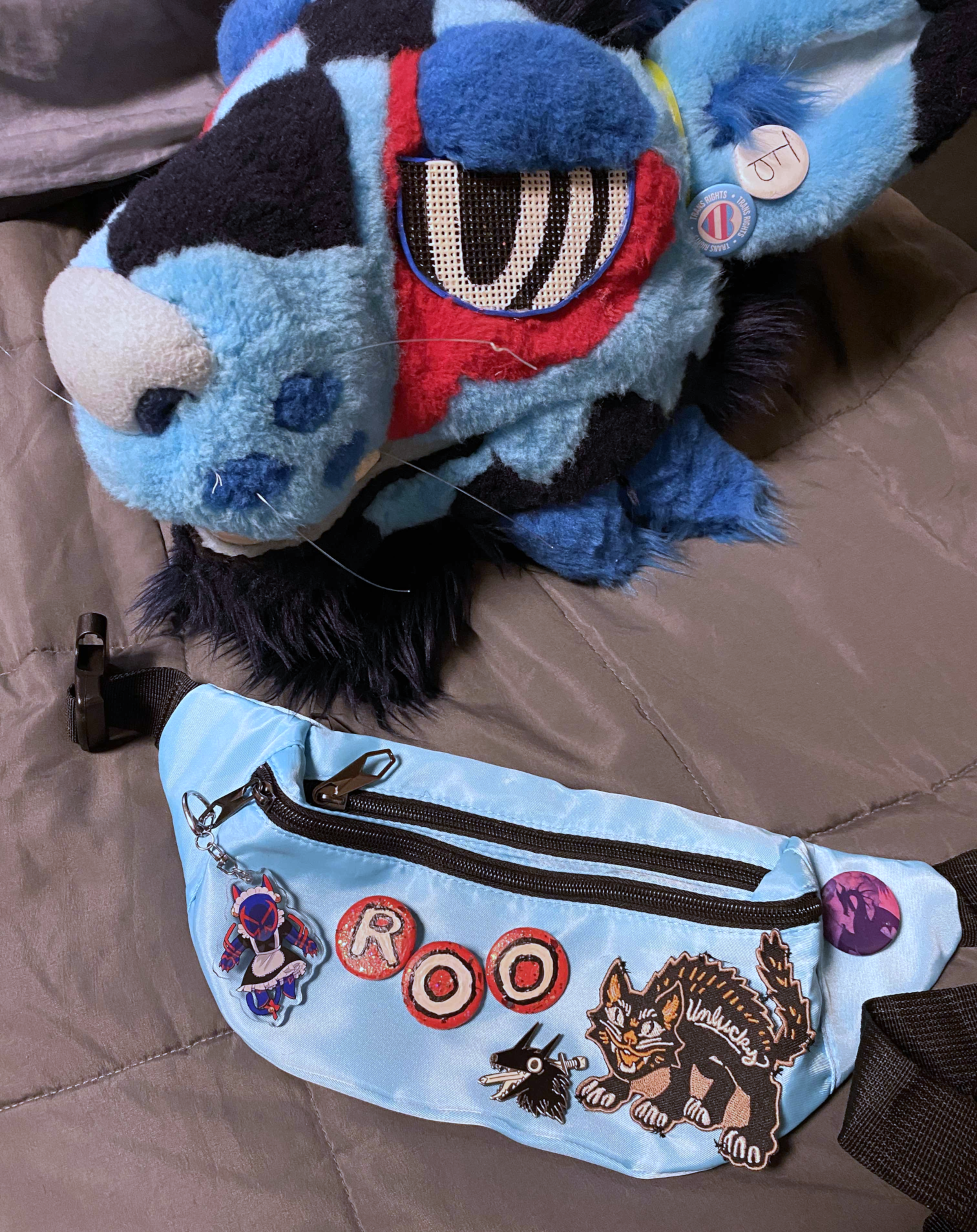 a blue kangaroo fursuit head laying on a gray weighted blanket. beneath it is an aqua fanny pack with three red pins on them, each one a letter that spells out "roo" as in "kangaroo"