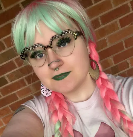 A real life photograph of me, a white feminine individual with a pink and mint green wig with braids on both sides, heart shaped glasses, and black lipstick.