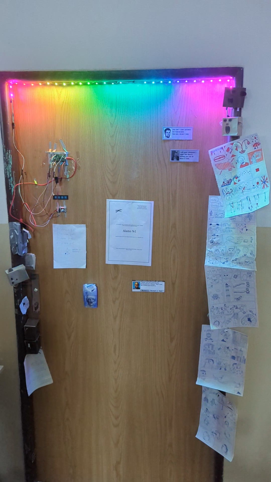 door with rgb led chain controlled by rasoverry pi pico, buttons and knobs duct taped to the door. there are also paper cubes with faces, ears and legs, my hand drawn comics, our teachers as video game characters in a dialog window saying silly things, pack of towels glued to the door, page torn out from a bible, and a parte for one of the cubes that my techer killed (torn apart).