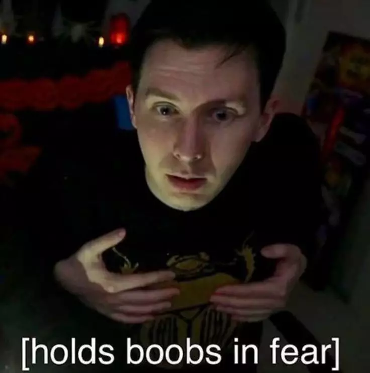 A white man with black hair and a black shirt on (with yellow writing); He is presumably a twitch streamer. The man stares lower than the camera, presumably at a computer monitor. The man is shocked, lips parted in fear, eyes wide; he probably reacts to what is on the monitor. The man has both of his hands cupped over each of his breasts. The caption of the image is [holds boobs in fear].